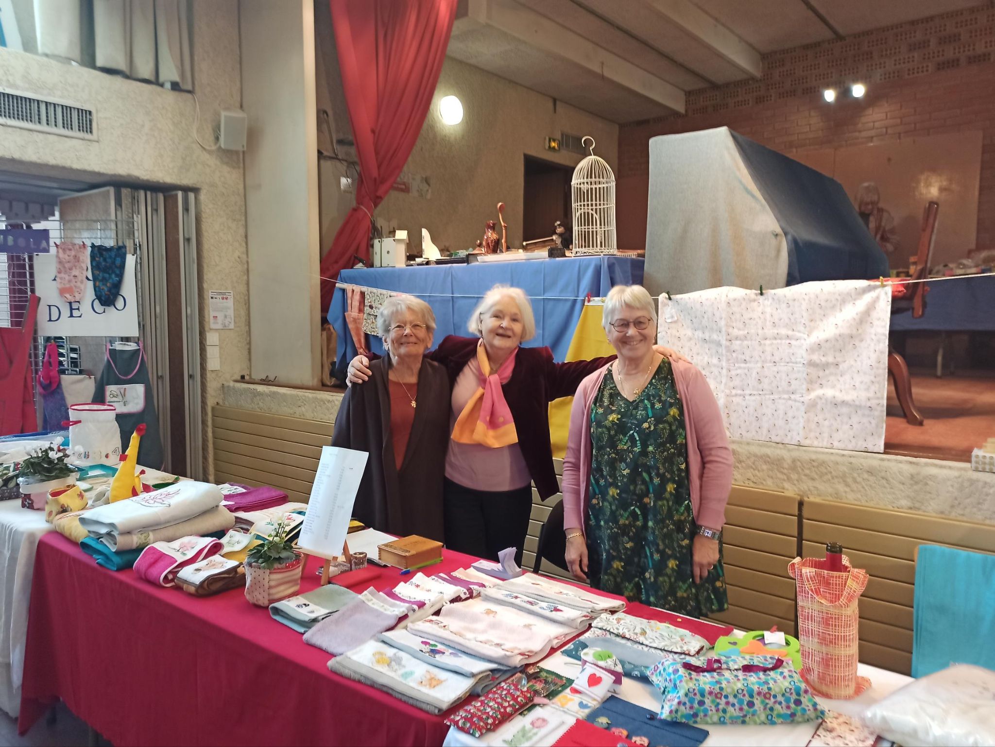 Trois bénévoles du groupe Art-Déco souriantes derrière leur stand, entourées de créations textiles brodées et cousues